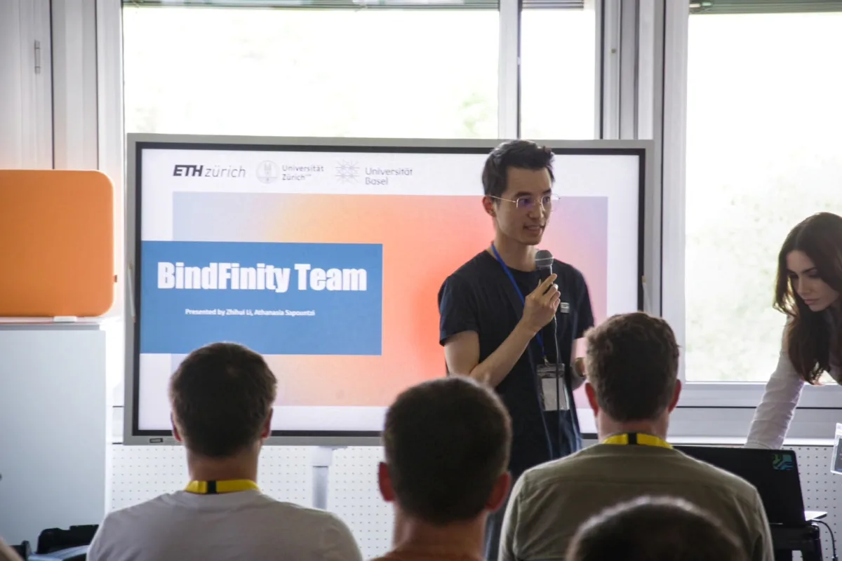 A speaker presenting to an audience beside a BindFinity team slide at ETH Zürich